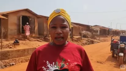 A women in re t-shirt and a yellow hair cover speaks, in the background you can see houses covered in red dust