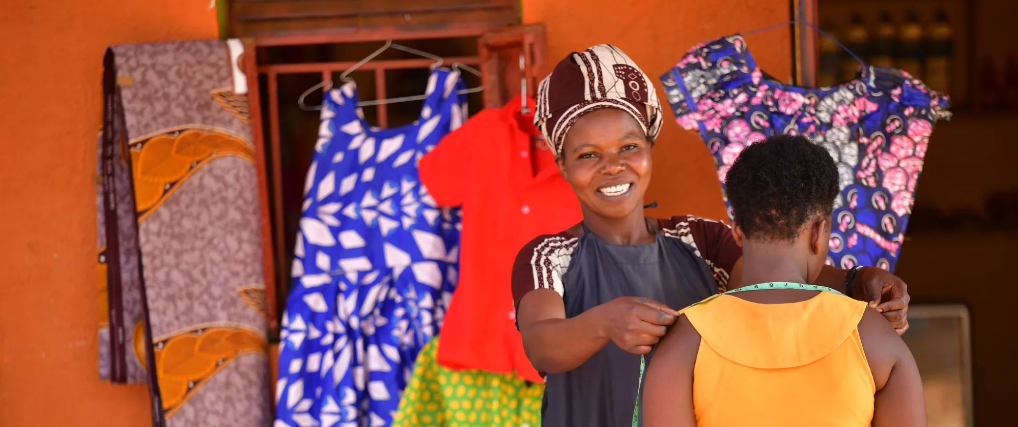 A female tailor smiles at the camera while she takes measurements of another woman's shoulders