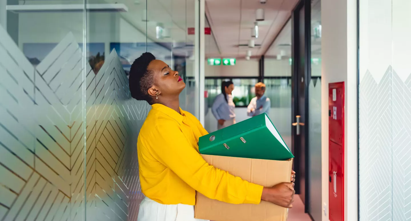 A young unhappy woman stands in a corridor holding a box of office supplies. 